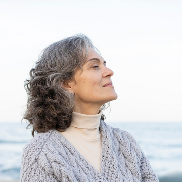 Woman with gray hair wearing a beige turtleneck and knitted cardigan against a blurred natural background
