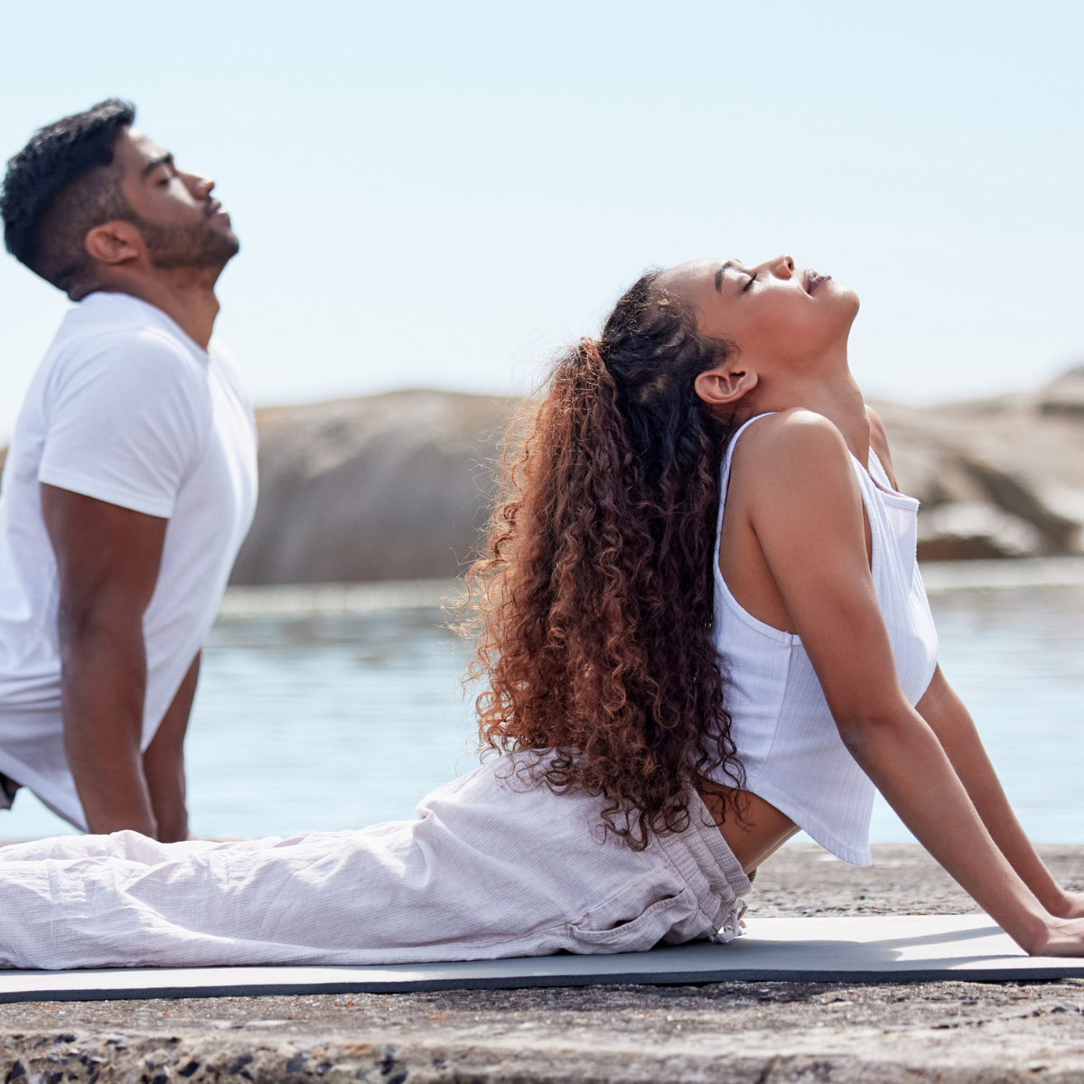 Man and woman sitting on a dock by water, both wearing white outfits.