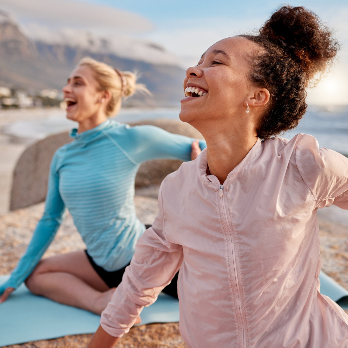 Two women laughing on yoga mats by a mountainous landscape