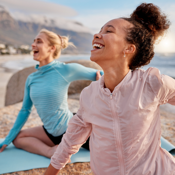 Two women laughing on yoga mats by a mountainous landscape