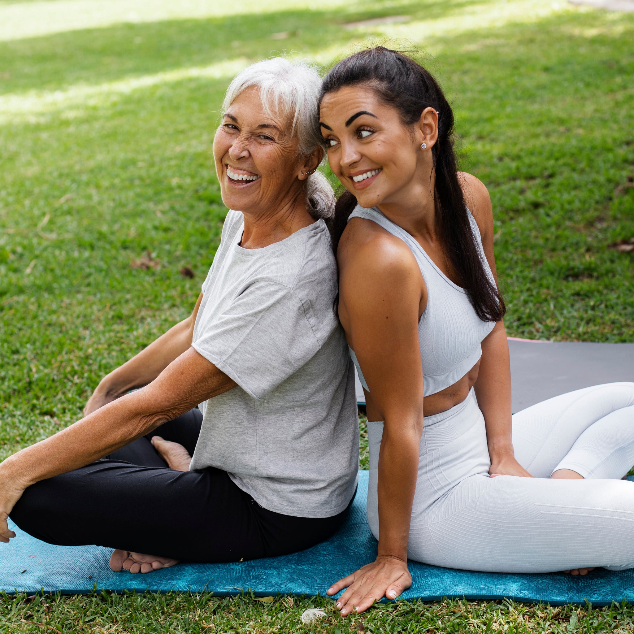 Two women sitting on a yoga mat outdoors, smiling.